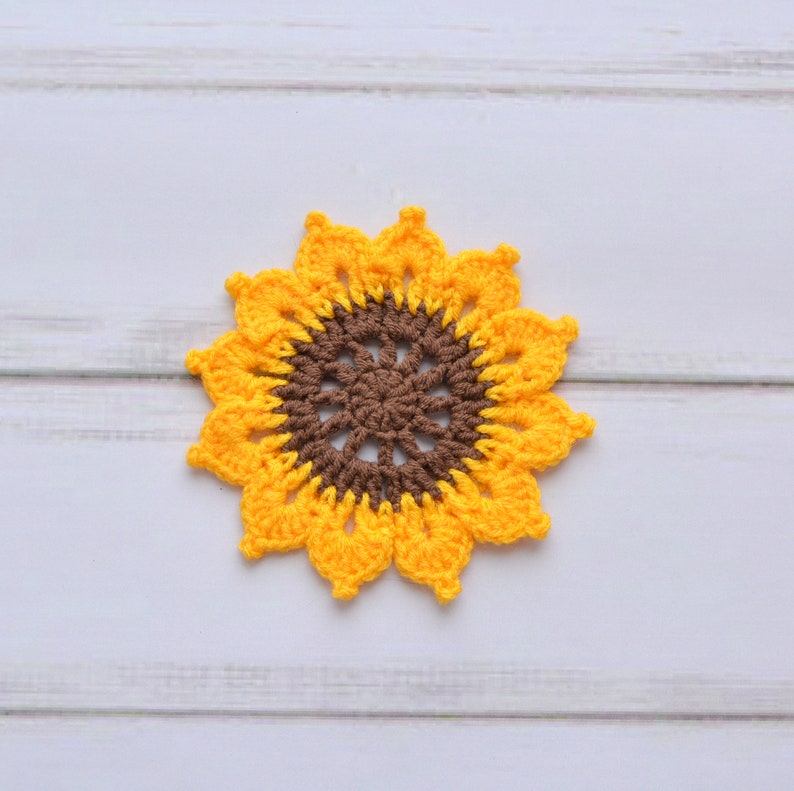 May include: A crocheted sunflower coaster in yellow and brown. The coaster is made of yarn and has a circular shape.