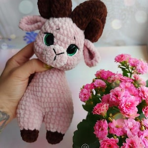 May include: A pink crocheted goat with brown horns and green eyes. The goat is holding a yellow flower in its mouth. The goat is sitting on a table with a pink flower pot in the background.