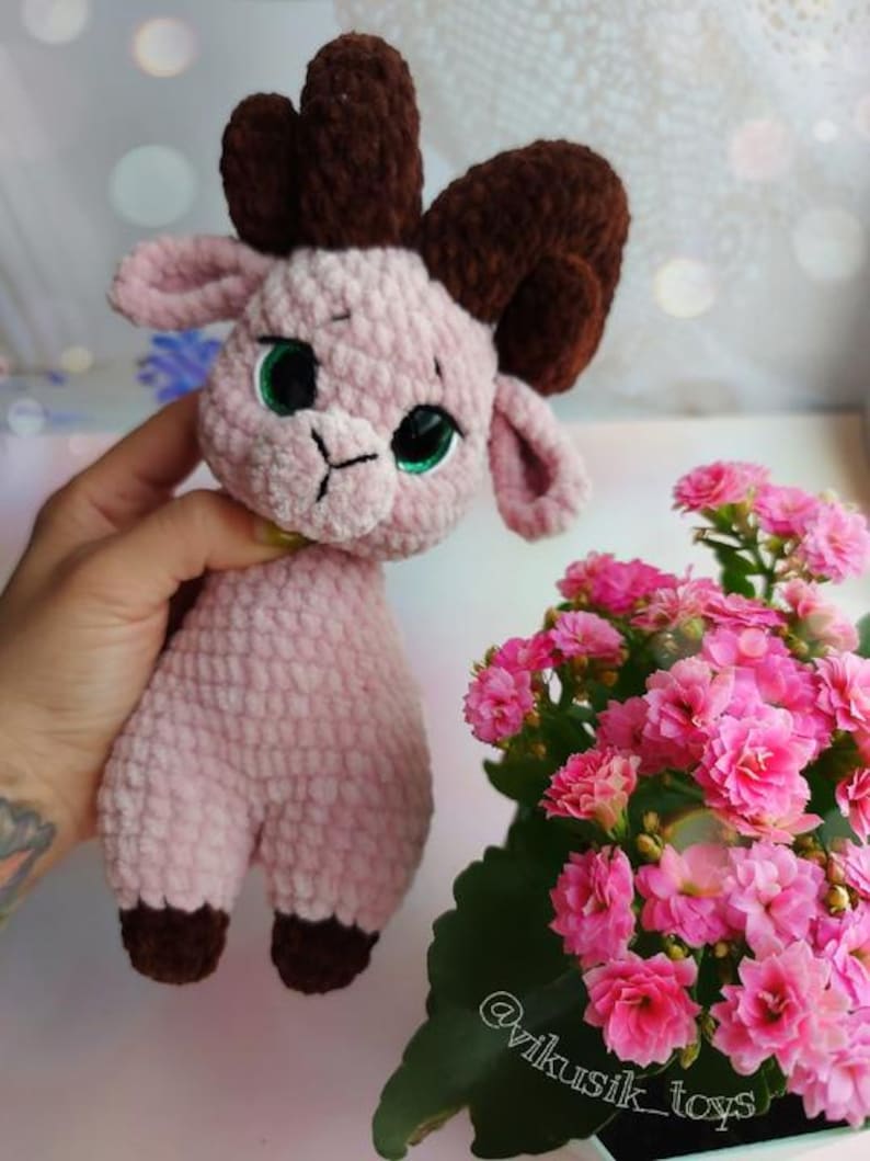May include: A pink crocheted goat with brown horns and green eyes. The goat is holding a yellow flower in its mouth. The goat is sitting on a table with a pink flower pot in the background.