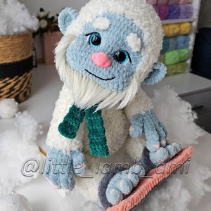 A white plush crochet yeti sits with its large blue feet resting on a pink and gray snowboard. Its fluffy body, long white beard, bright blue eyes, and dark green scarf create a playful winter sports character.