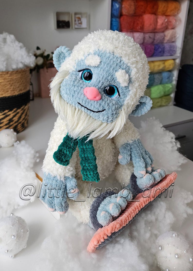 A white plush crochet yeti sits with its large blue feet resting on a pink and gray snowboard. Its fluffy body, long white beard, bright blue eyes, and dark green scarf create a playful winter sports character.