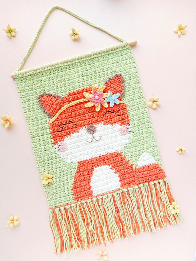 Op de afbeelding: Een gehaakte wandhanger met een vos met een bloemenkrans. De vos is oranje met witte accenten en is geplaatst tegen een groene achtergrond. De hanger heeft een franje van groen en oranje garen.