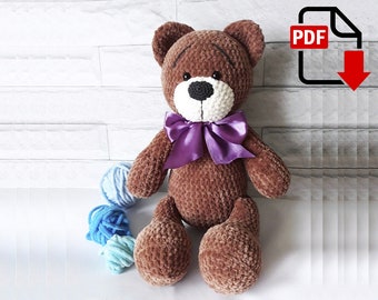 Könnte beinhalten: Ein brauner gehäkelter Teddybär mit einer violetten Schleife. Der Bär sitzt auf einer weißen Oberfläche mit blauen und weißen Wollknäueln.