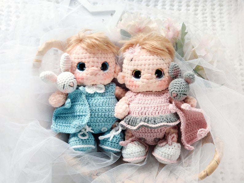 Pu&ograve; includere: Due bambole all'uncinetto, una in blu e una in rosa, tengono in braccio dei compagni di peluche. Le bambole indossano abiti all'uncinetto e hanno tratti del viso ricamati.