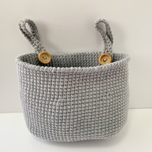 May include: A gray crocheted hanging basket with two wooden button handles. The basket is made with a textured stitch and has a rounded shape.
