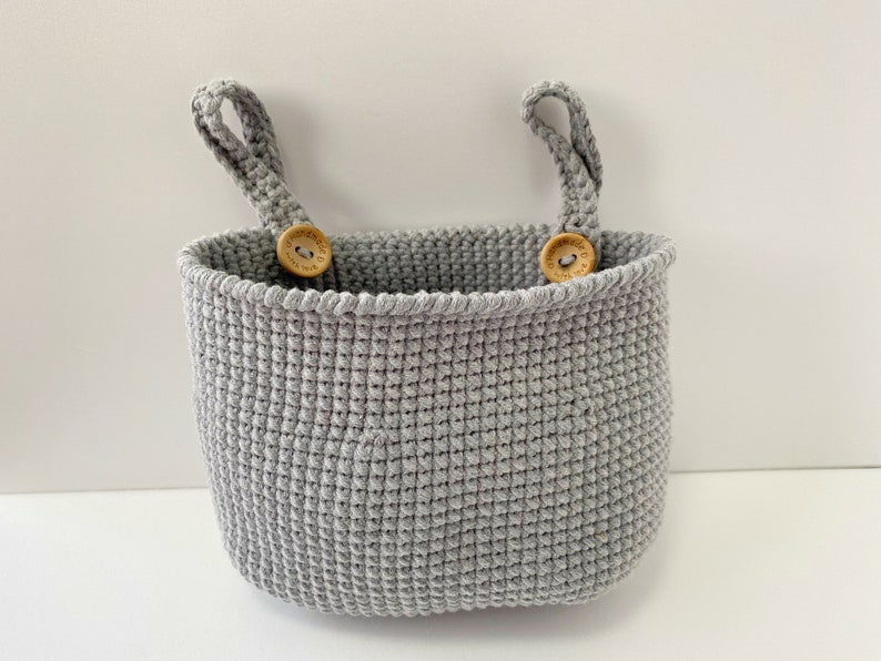 May include: A gray crocheted hanging basket with two wooden button handles. The basket is made with a textured stitch and has a rounded shape.
