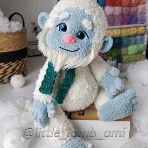 A seated crochet yeti made from soft white fluffy yarn has oversized blue feet, pink toenails, and a smiling face with bright blue eyes. A dark green scarf hangs around its neck, adding contrast to the winter-themed plush toy.