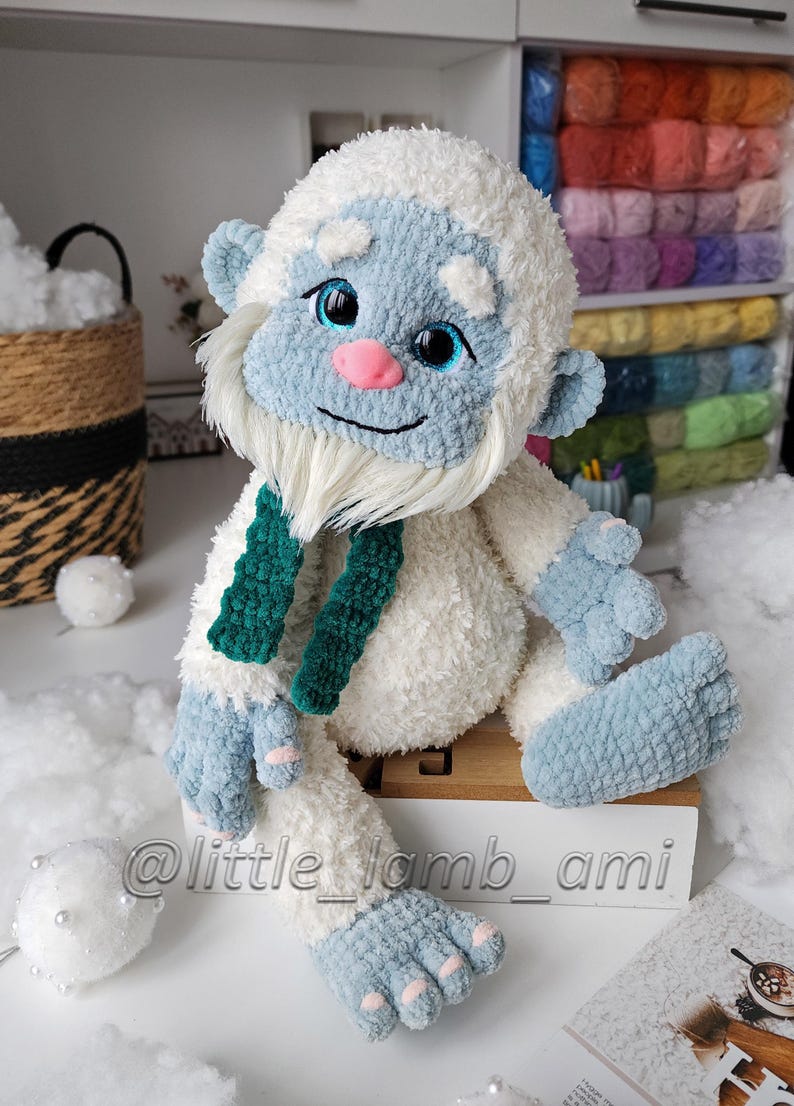 A seated crochet yeti made from soft white fluffy yarn has oversized blue feet, pink toenails, and a smiling face with bright blue eyes. A dark green scarf hangs around its neck, adding contrast to the winter-themed plush toy.