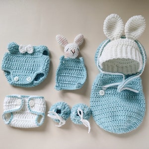 Pu&ograve; includere: Un set di vestiti per neonati all'uncinetto in blu e bianco, tra cui un sacco a pelo con cappuccio, un body, un pannolino, scarpine e un sonaglio a forma di coniglio.