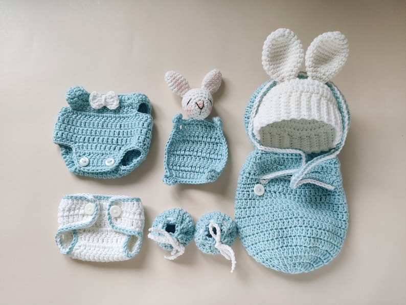 Pu&ograve; includere: Un set di vestiti per neonati all'uncinetto in blu e bianco, tra cui un sacco a pelo con cappuccio, un body, un pannolino, scarpine e un sonaglio a forma di coniglio.