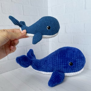 May include: Two blue crocheted whale plushies. The smaller whale is held in a hand. The larger whale is lying on a white surface.