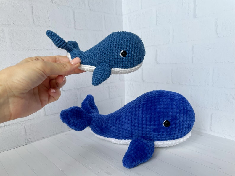 May include: Two blue crocheted whale plushies. The smaller whale is held in a hand. The larger whale is lying on a white surface.