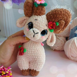 May include: A crocheted plush toy lamb with brown horns and pink flowers. The lamb is light pink and has a sweet expression. The toy is handmade and has a unique, whimsical style.