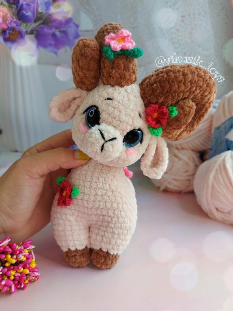 May include: A crocheted plush toy lamb with brown horns and pink flowers. The lamb is light pink and has a sweet expression. The toy is handmade and has a unique, whimsical style.