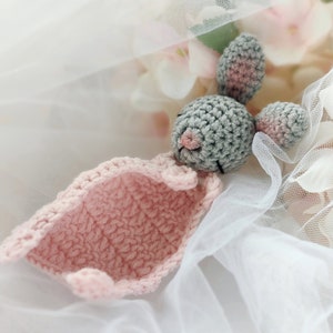 Pu&ograve; includere: Un coniglietto di uncinetto grigio con orecchie rosa e una coperta di uncinetto rosa. Il coniglietto &egrave; sdraiato su un tessuto di tulle bianco.