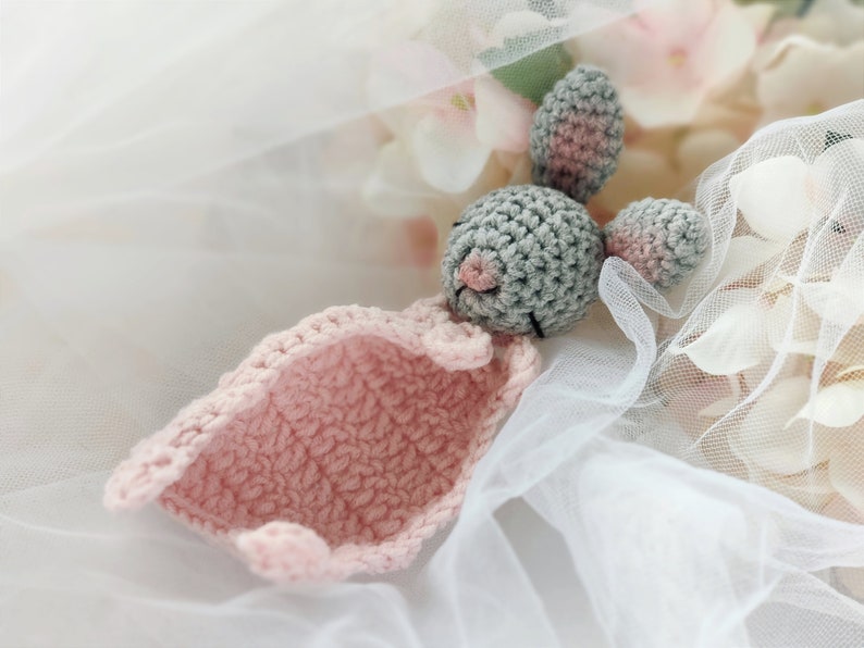 Pu&ograve; includere: Un coniglietto di uncinetto grigio con orecchie rosa e una coperta di uncinetto rosa. Il coniglietto &egrave; sdraiato su un tessuto di tulle bianco.