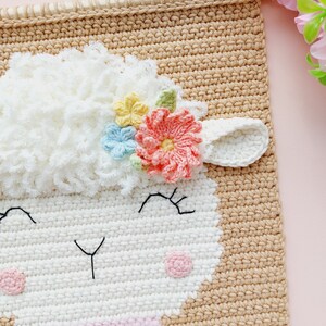 May include: A crocheted wall hanging featuring a white sheep with a pink, yellow, and blue flower crown. The sheep has a pink nose and black eyes. The background is a light brown.