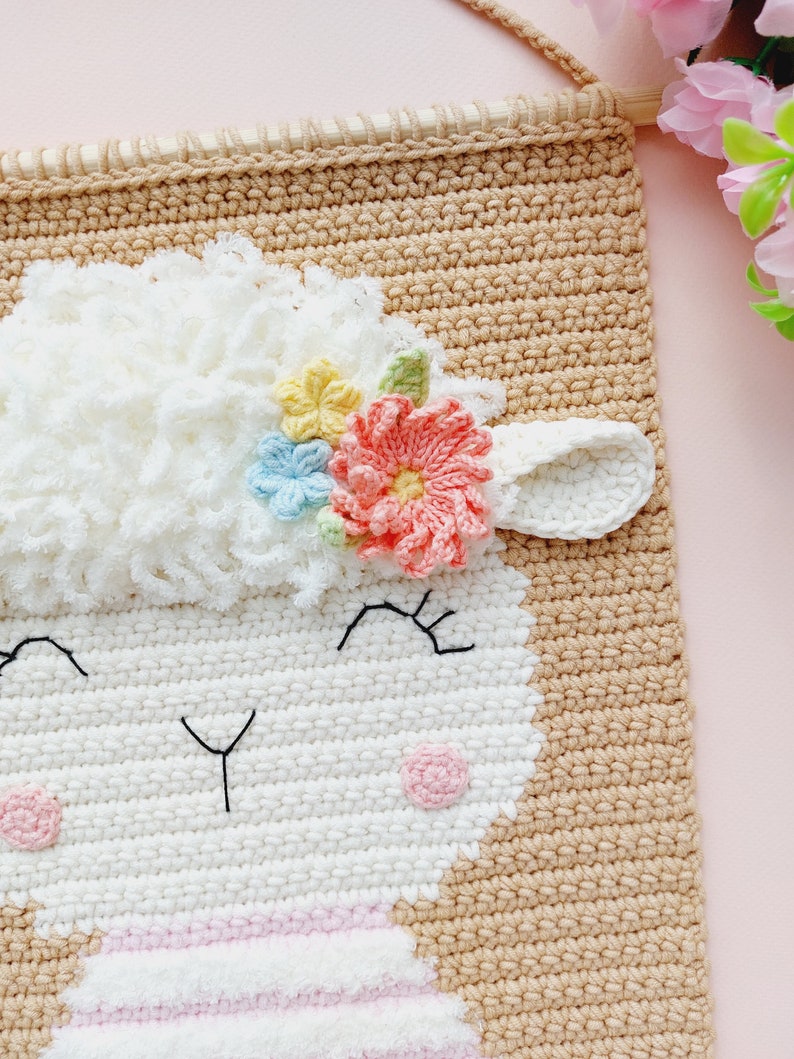 May include: A crocheted wall hanging featuring a white sheep with a pink, yellow, and blue flower crown. The sheep has a pink nose and black eyes. The background is a light brown.
