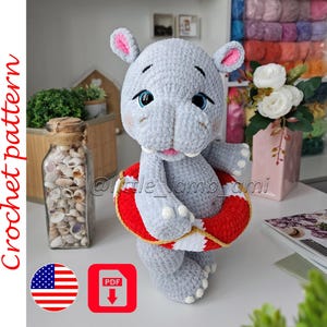 May include: A grey crocheted hippo plush toy with large blue eyes, pink inner ears, and rosy cheeks. The hippo is holding a red and white striped life preserver ring. The text Crochet pattern is visible on the left side of the image.