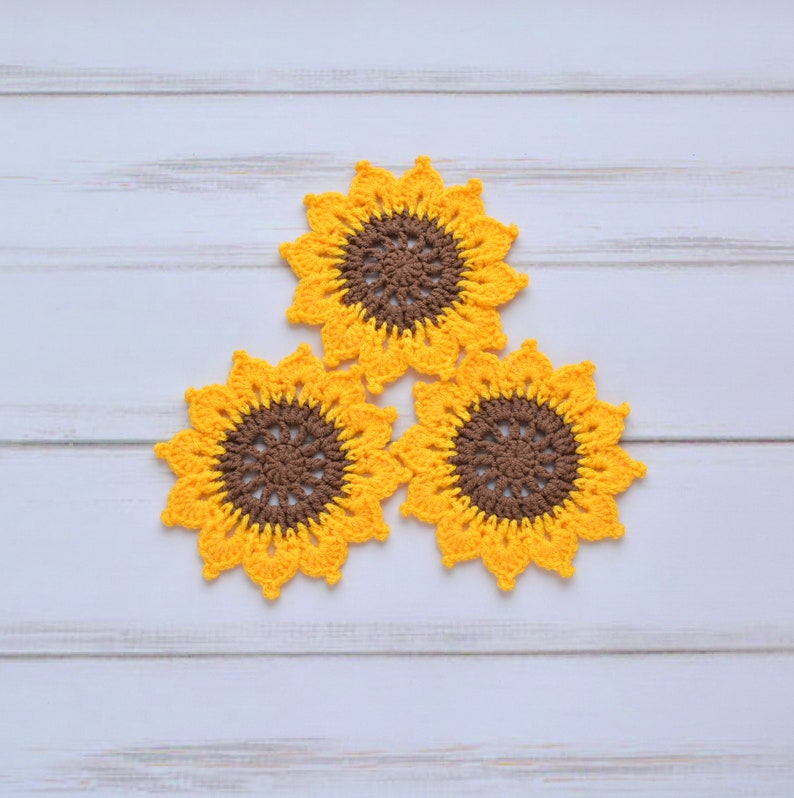 May include: Three crocheted sunflower coasters in yellow and brown. The coasters are arranged in a triangle shape on a white wooden surface.