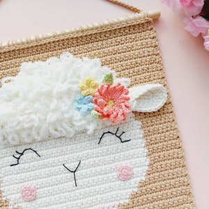 May include: A crocheted wall hanging featuring a white sheep with a pink, yellow, and blue flower crown. The sheep has a pink nose and black eyes. The wall hanging is made with beige yarn and has a wooden dowel at the top.