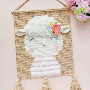 May include: A crocheted wall hanging featuring a white lamb with pink and white stripes, a fluffy white head, and a flower crown with pink, blue, yellow, and green flowers. The hanging has three beige tassels.