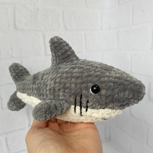 Puede incluir: Un juguete de peluche de tibur&oacute;n gris tejido a crochet con detalles en fieltro blanco alrededor de la boca y un ojo bordado en negro.
