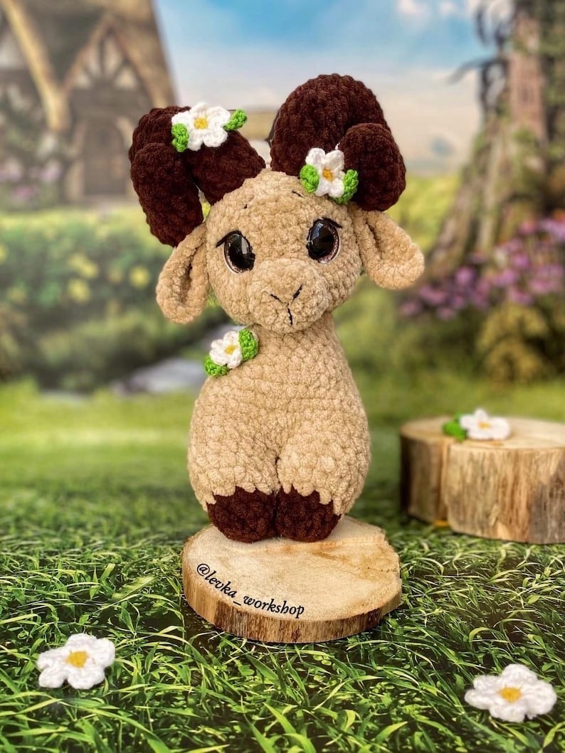 May include: A crocheted plush toy ram with brown horns and a beige body. The ram has large black eyes and is decorated with white flowers. The toy is sitting on a wooden slice with the text "@leoka_workshop" written on it.