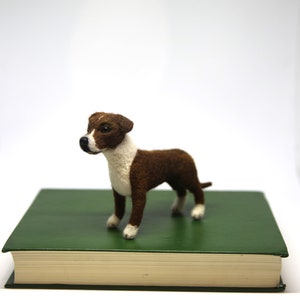 May include: A handcrafted felt dog figurine in brown and white stands on a green book. The dog has a white chest, face, and paws, with brown fur on its back and head. The background is plain white.