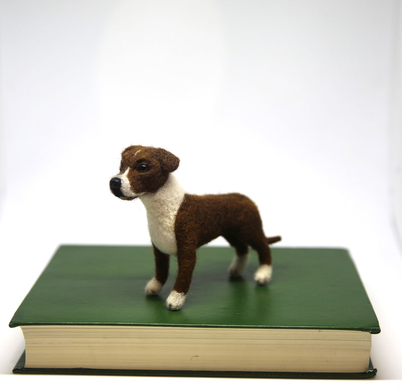 May include: A handcrafted felt dog figurine in brown and white stands on a green book. The dog has a white chest, face, and paws, with brown fur on its back and head. The background is plain white.