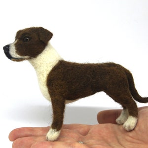 May include: A handcrafted felt dog figurine in a standing pose. The dog has brown fur with white markings on its chest, neck, and paws. The dog's head is brown with white and black details. The figurine is held in a person's hand.