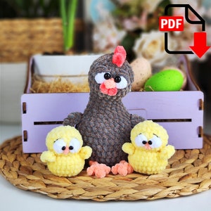 May include: A handmade crochet chicken and two chicks sit on a woven mat. The grey chicken has a red comb and beak, with two yellow chicks. A purple wooden crate with Easter eggs is in the background.