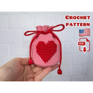 May include: A pink crocheted drawstring bag with a red heart design. The bag has a red drawstring closure and red square beads at the ends. The image also includes the text "CROCHET PATTERN", a US flag, and a PDF icon.