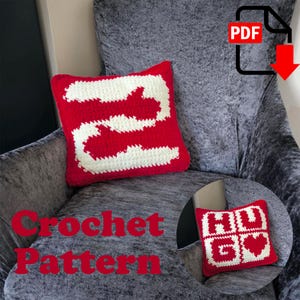 May include: A red and white crochet pillow with a large white number 2 design. The pillow is square and sits on a gray armchair. The words "Crochet Pattern" are written in red across the bottom of the image.