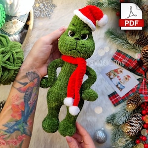 Puede incluir: Personaje de Grinch de crochet verde con un gorro de Papá Noel rojo y blanco y una bufanda roja con pompones blancos. El Grinch tiene una expresión gruñona. La imagen incluye el texto "@VIKUSIK_TOYS".