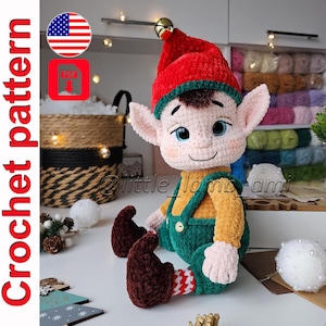 Op de afbeelding: Gehaakte elfenpop met een rode hoed, groene tuinbroek en bruine schoenen. De pop heeft een lachend gezicht met blauwe ogen en roze wangen. De afbeelding bevat de tekst "Crochet pattern" en een US vlag icoon.