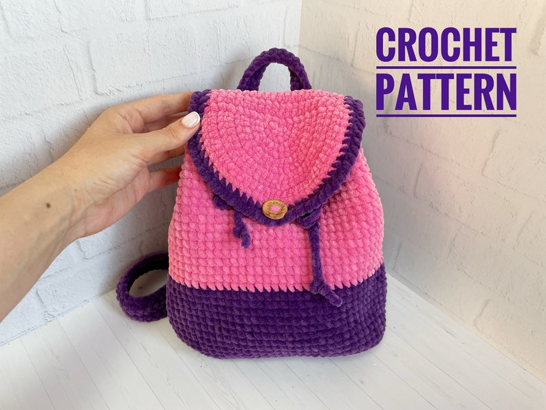 Pu&ograve; includere: Uno zaino in crochet rosa e viola con chiusura a bottone marrone. CROCHET PATTERN &egrave; scritto in testo viola nella parte superiore dell'immagine.