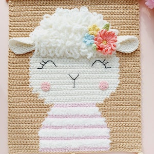 May include: A crocheted wall hanging featuring a white lamb with pink and white stripes, a fluffy white head, and a flower crown with pink, blue, and yellow flowers. The lamb has black eyes and a black nose. The wall hanging is beige and has two tassels.