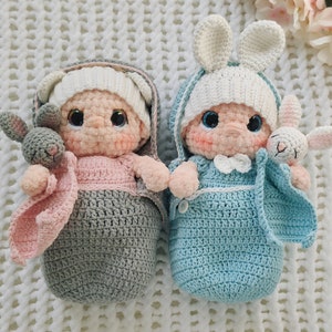 Pu&ograve; includere: Due bambole per bambini all'uncinetto, una in un sacco a pelo grigio e rosa con un coniglietto grigio, e l'altra in un sacco a pelo blu con un coniglietto bianco.