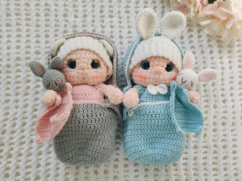 Pu&ograve; includere: Due bambole per bambini all'uncinetto, una in un sacco a pelo grigio e rosa con un coniglietto grigio, e l'altra in un sacco a pelo blu con un coniglietto bianco.