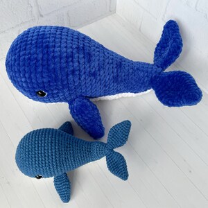 May include: Two crocheted whale plushies, one large and one small, both blue with white accents. The larger whale is lying on its side with its tail curled up. The smaller whale is standing upright with its tail behind it.