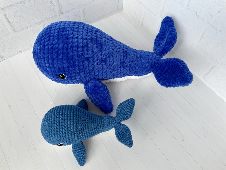 May include: Two crocheted whale plushies, one large and one small, both blue with white accents. The larger whale is lying on its side with its tail curled up. The smaller whale is standing upright with its tail behind it.