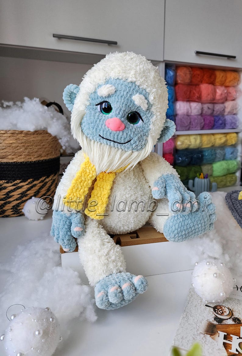以下が含まれることがあります： 白いふわふわした毛並み、水色の顔、手、足、そして鮮やかな黄色のスカーフを身につけたイエティのぬいぐるみ。大きな暗い目、ピンクの鼻、白いあごひげが特徴。中央には「@little_lamb_ami」というテキストが表示されている。