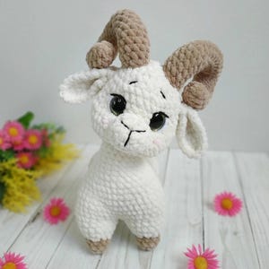 May include: A white crocheted goat with brown crocheted horns. The goat has large black eyes and a black stitched nose and mouth. The goat is standing on a white wooden surface with pink flowers.