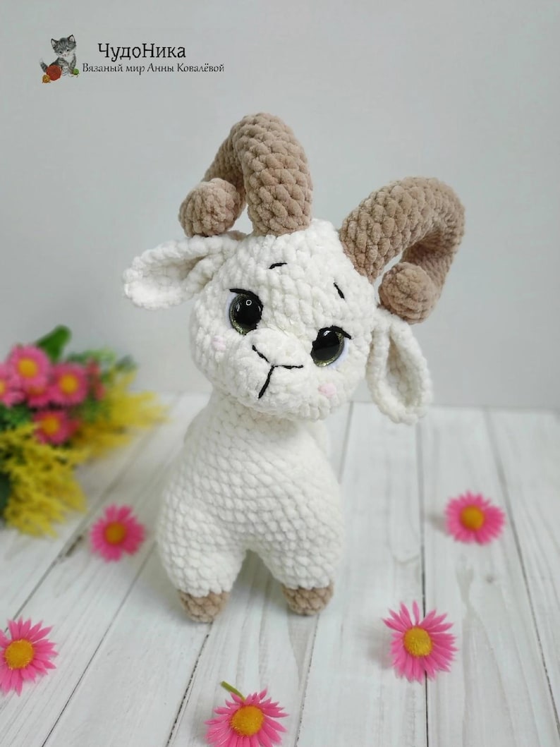 May include: A white crocheted goat with brown crocheted horns. The goat has large black eyes and a black stitched nose and mouth. The goat is standing on a white wooden surface with pink flowers.