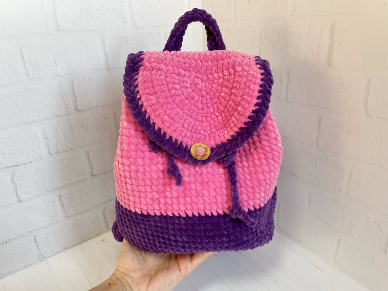 Pu&ograve; includere: Uno zaino in crochet rosa e viola con chiusura a bottone in legno. Lo zaino ha una chiusura a coulisse ed &egrave; realizzato con un filato morbido e soffice.