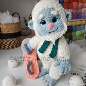 A fluffy white crochet yeti with blue hands and feet, a pink nose, and a long white beard stands holding a pink and gray snowboard. The plush amigurumi toy wears a dark green scarf, with colorful yarn shelves in the background.