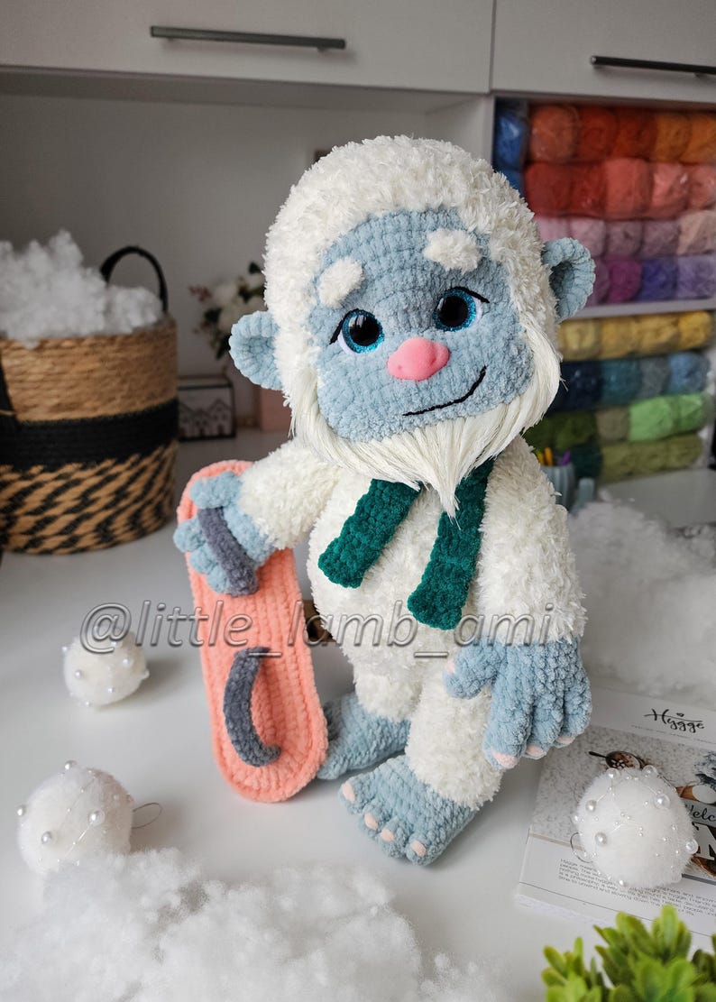 A fluffy white crochet yeti with blue hands and feet, a pink nose, and a long white beard stands holding a pink and gray snowboard. The plush amigurumi toy wears a dark green scarf, with colorful yarn shelves in the background.