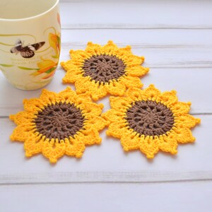 May include: Three crocheted sunflower coasters in yellow and brown. The coasters are on a white wooden surface.