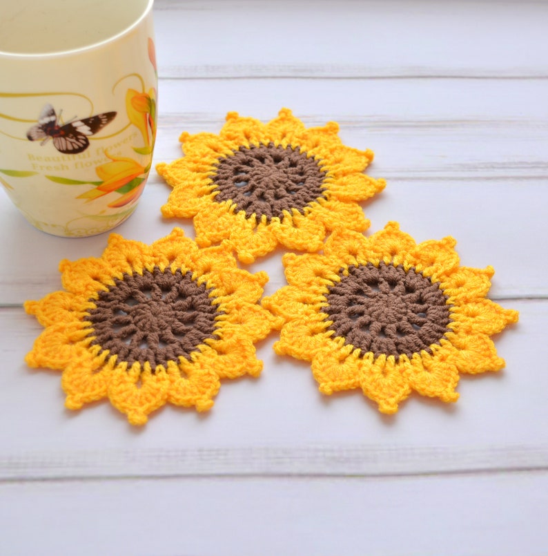 May include: Three crocheted sunflower coasters in yellow and brown. The coasters are on a white wooden surface.